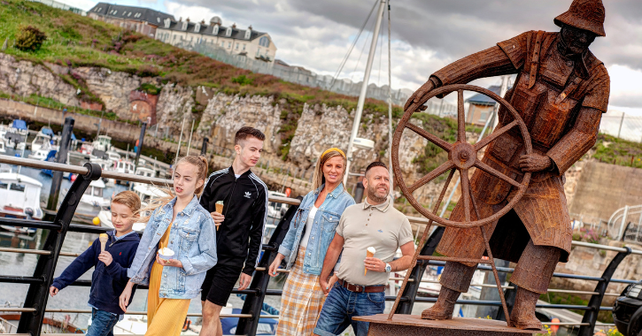 Family walking eating ice cream at Seaham Marina with sculpture of The Coxwain on display.